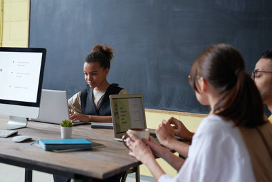 three people on devices in a classroom with a chalkboard in the background